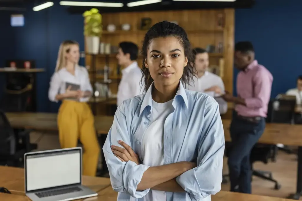 Mulher empreendedora de um dos tipos de empreendedorismo com braços cruzados à frente com colegas de trabalho ao fundo tendo conversas informais.